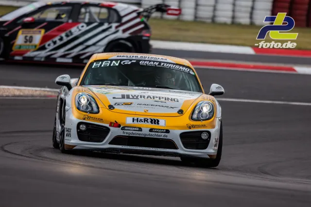 Porsche Cayman in orange-weißer Lackierung auf der Nordschleife beim Qualifying, im Hintergrund ein weiteres Fahrzeug.
