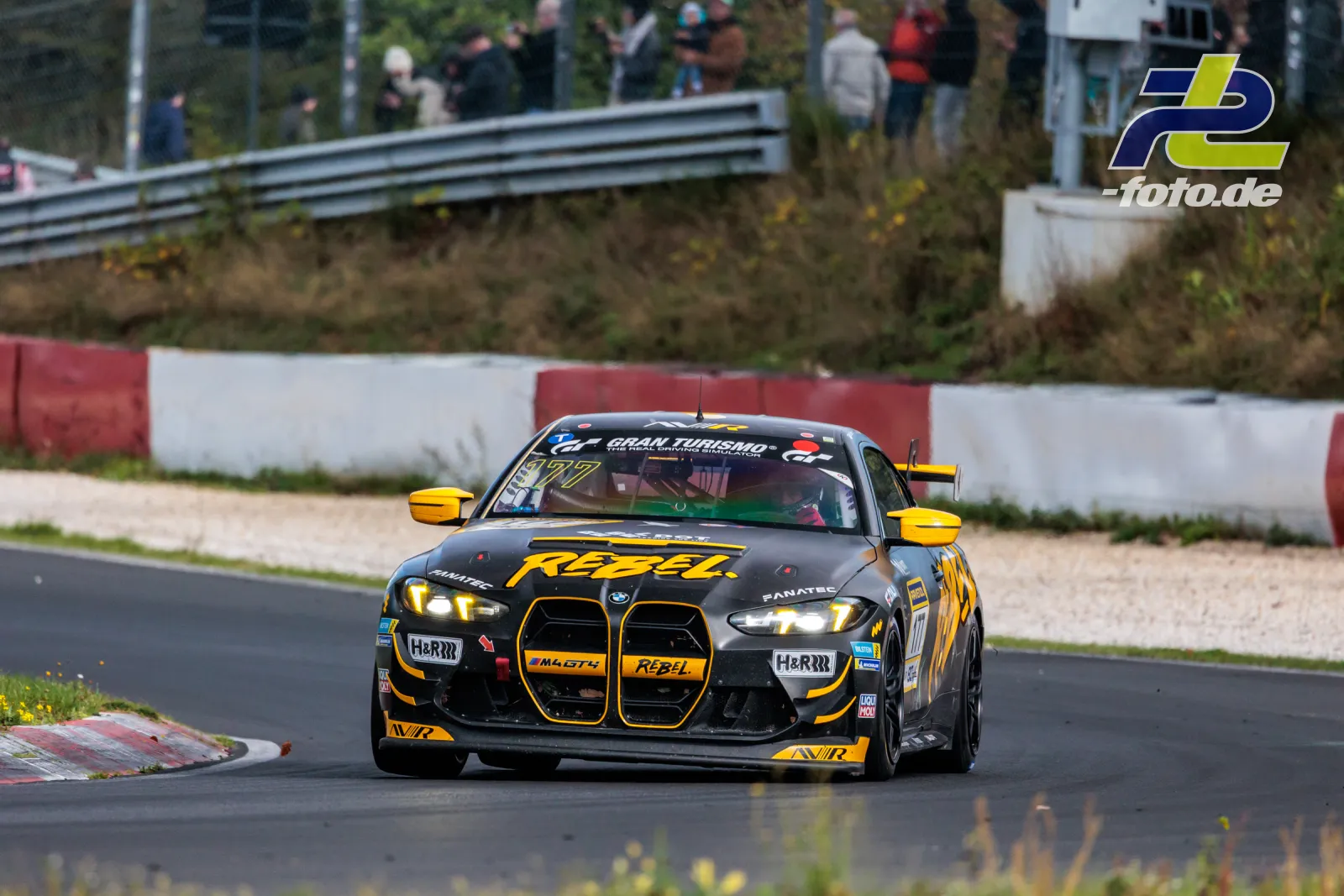 BMW M4 GT4 #177 in schwarz-gelber RËBEL-Lackierung in einer Kurve auf dem Nürburgring mit Zuschauern im Hintergrund.