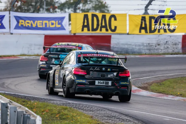 Schwarzer BMW M235i Racing auf der Nürburgring-Nordschleife, verfolgt von einem weiteren Fahrzeug, ADAC-Werbebanden im Hintergrund.
