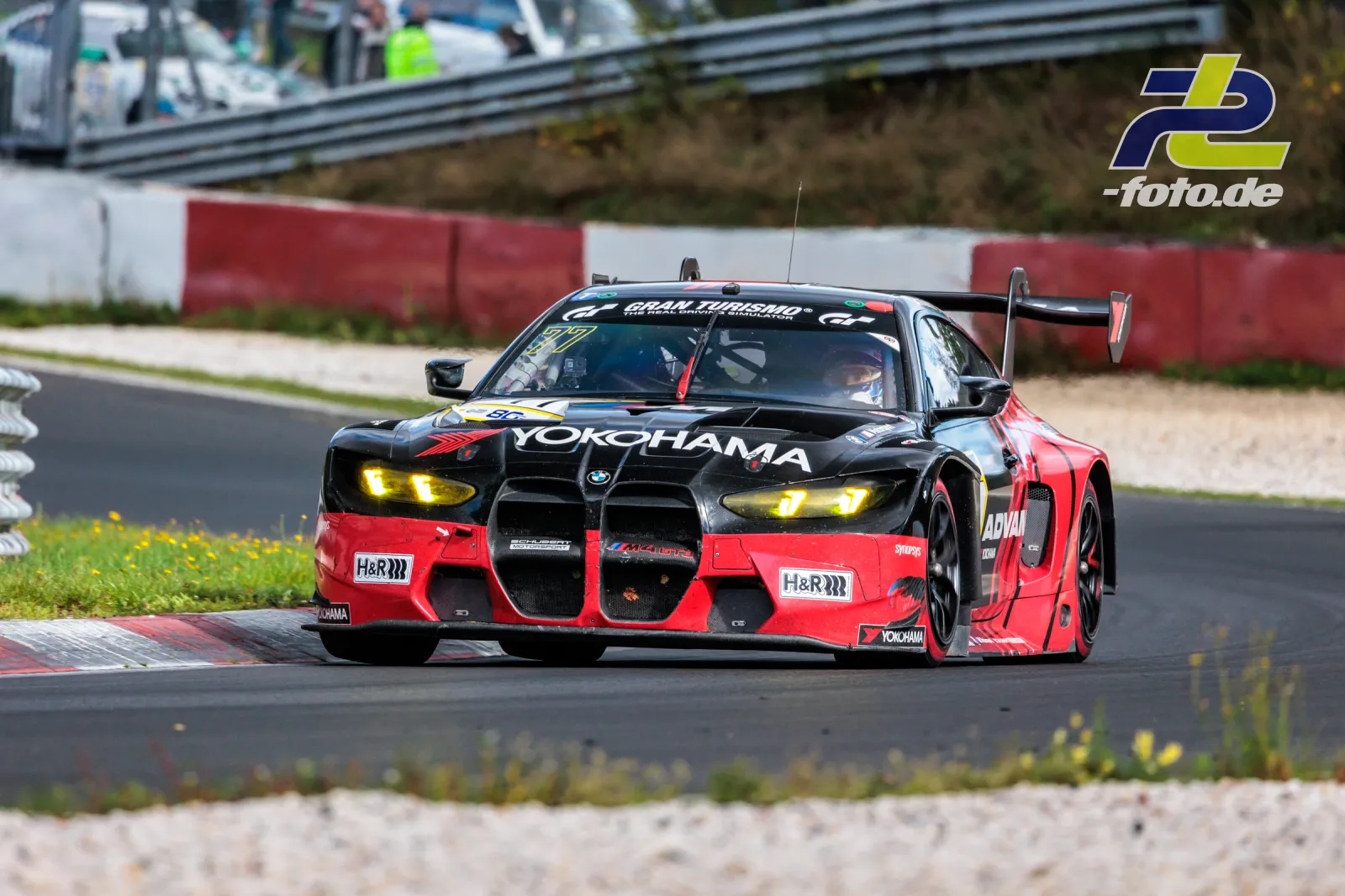 BMW M4 GT3 in rot-schwarzer Yokohama-Lackierung auf der Nordschleife beim Rennen, Startnummer 77, gelbe Scheinwerfer eingeschaltet.