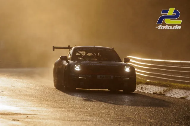Porsche 911 GT3 R im Gegenlicht bei der Ausfahrt aus einer Kurve, Scheinwerfer eingeschaltet, Staub aufwirbelnd.