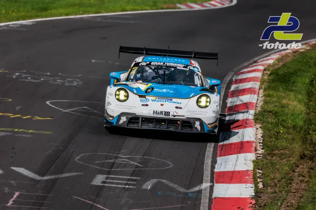 Porsche 911 GT3 R in hellblau-weißer Lackierung fährt durch eine Kurve auf dem Nürburgring. Rot-weiße Randsteine und Wiese im Hintergrund.