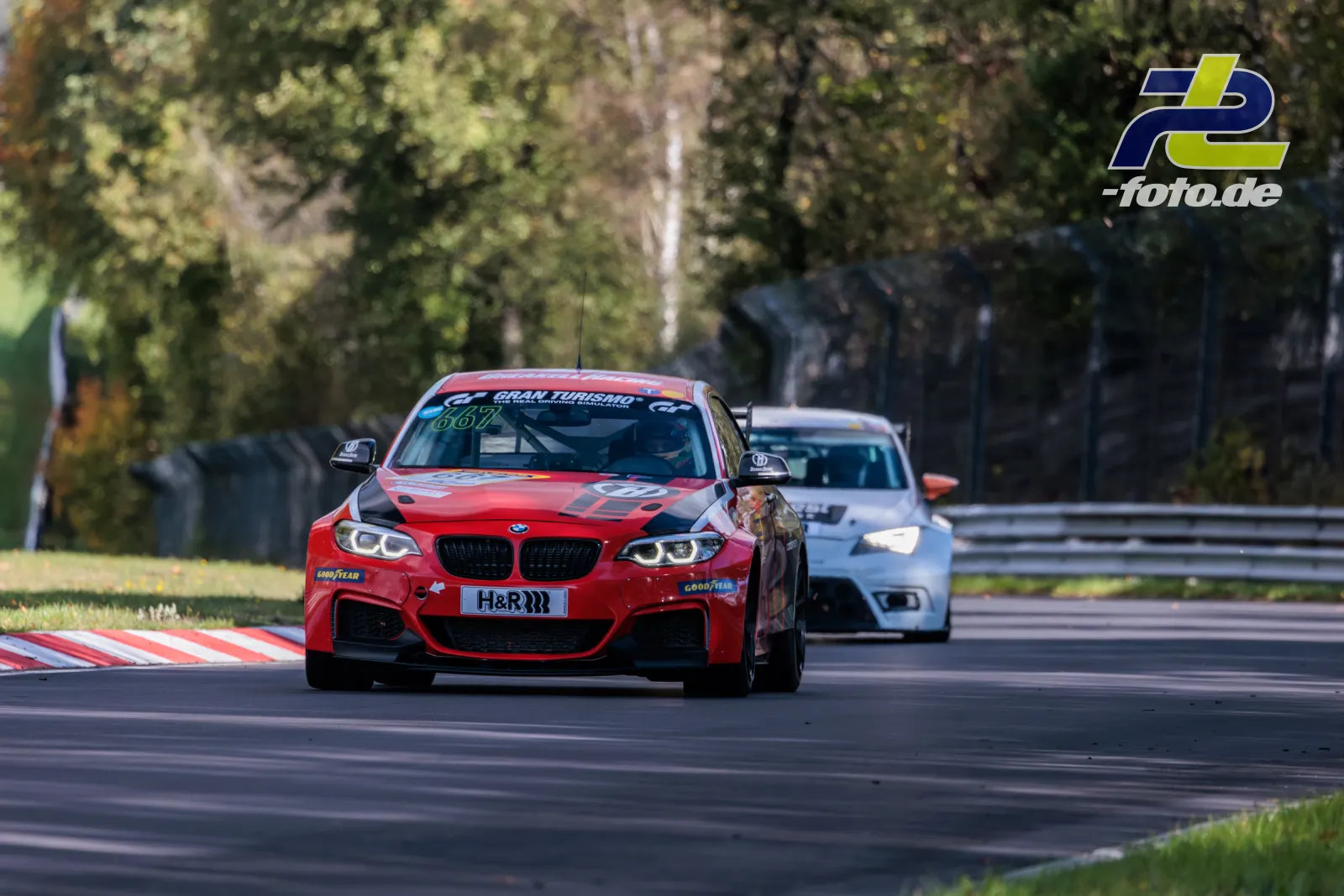 Roter BMW M240i #667 führt vor weißem BMW auf der Nordschleife beim NLS-Rennen, im Hintergrund Bäume und Streckenbegrenzung.