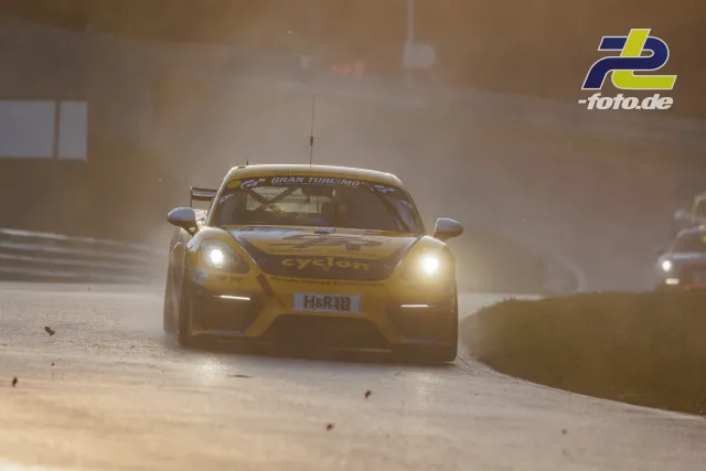 Gelber Porsche Cayman mit Cyclon-Sponsoring auf der Nordschleife in staubiger Atmosphäre, Frontansicht mit eingeschalteten Scheinwerfern.