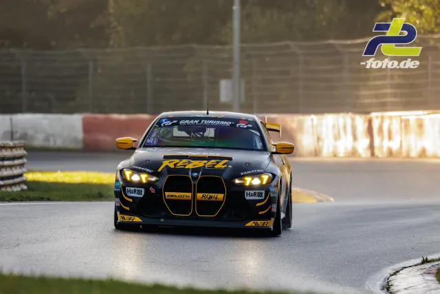 BMW M4 GT3 in schwarz-gelber REBEL-Lackierung auf dem Nürburgring beim Qualifying, Frontansicht in Kurve mit Gegenlicht.