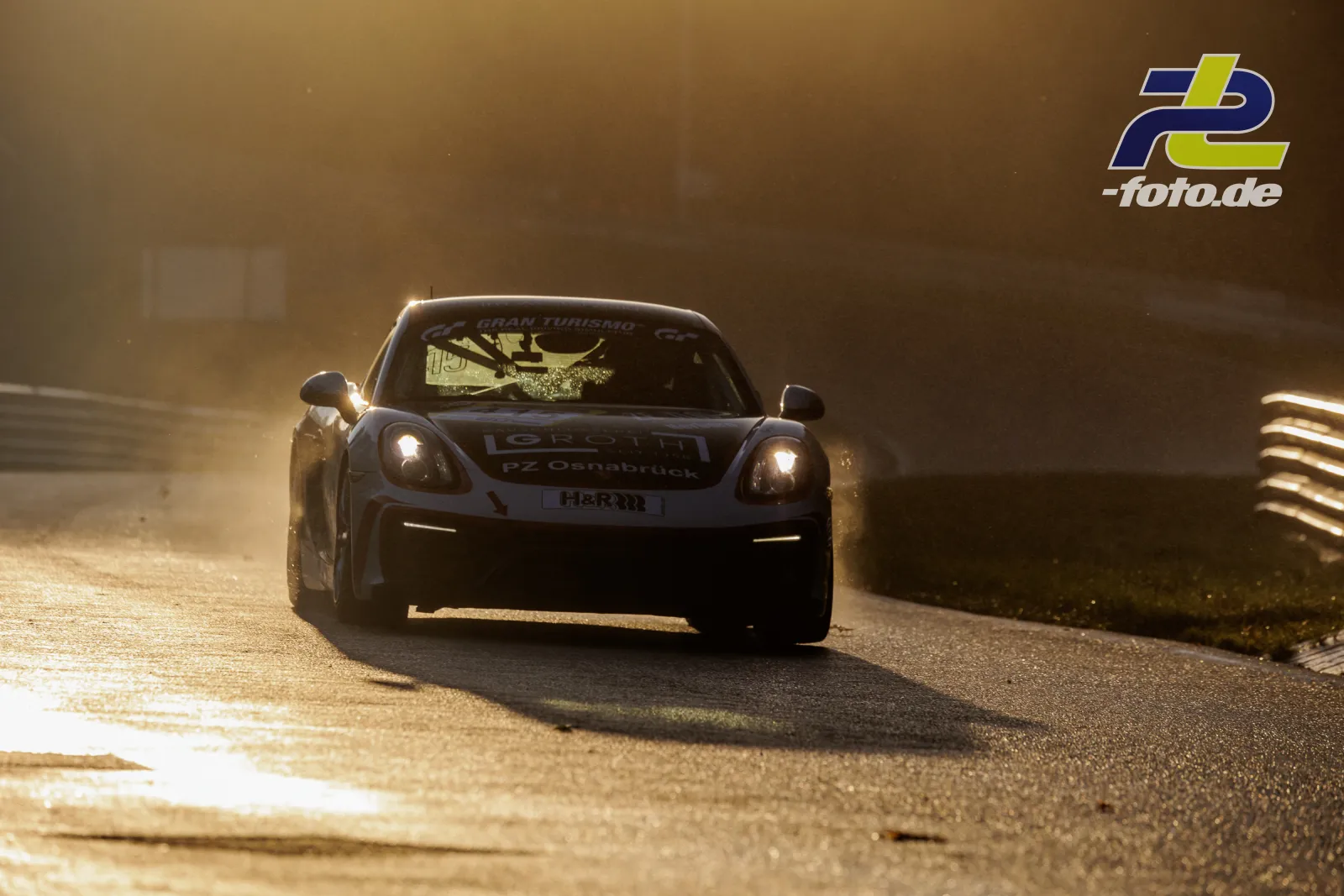 Porsche Cayman S im Gegenlicht auf dem Nürburgring während des Qualifyings zur NLS Sportwarte-Trophy, Frontalansicht mit eingeschalteten Scheinwerfern.