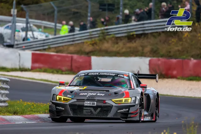 Audi R8 LMS GT3 Nummer 50 in grau-roter Lackierung mit gelben Scheinwerfern auf der Nordschleife, Zuschauer im Hintergrund.