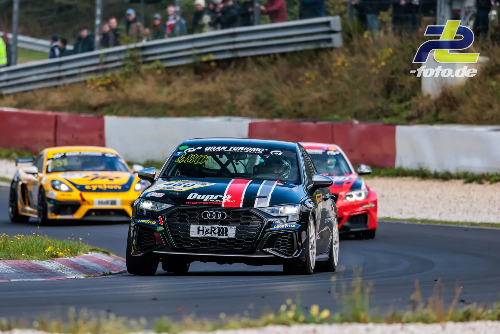 Drei Rennwagen in enger Folge auf der Nordschleife, vorne ein Audi mit rot-weißen Streifen, gefolgt von einem gelben und einem roten Auto.