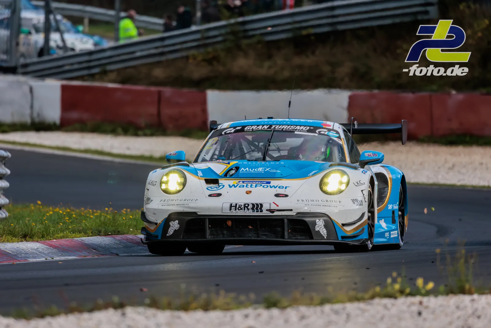 Porsche 911 GT3 R in Weiß-Blauer Watt Power Lackierung in der Nordschleifenkurve mit leuchtenden Scheinwerfern