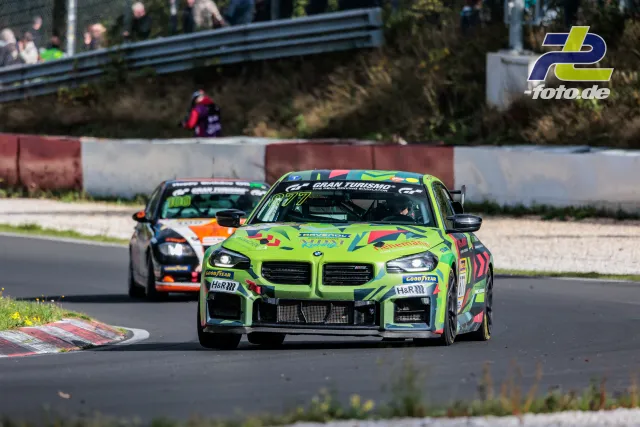 Zwei Rennwagen im Zweikampf auf der Nordschleife beim NLS-Rennen, vorne ein neongrüner BMW M4 GT4, gefolgt von einem schwarz-orangen Fahrzeug.