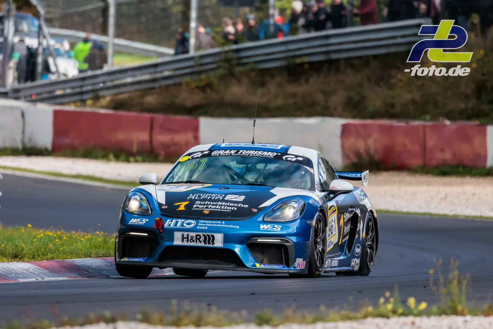 Blauer Porsche 718 Cayman GT4 #961 mit Hühr-Sponsoring auf der Nordschleife beim NLS-Rennen, Zuschauer im Hintergrund.