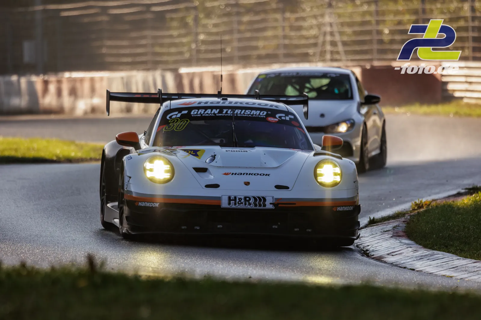 Porsche 911 GT3 R #30 in weiß-oranger Lackierung fährt durch eine Kurve am Nürburgring, gefolgt von einem weiteren Fahrzeug.