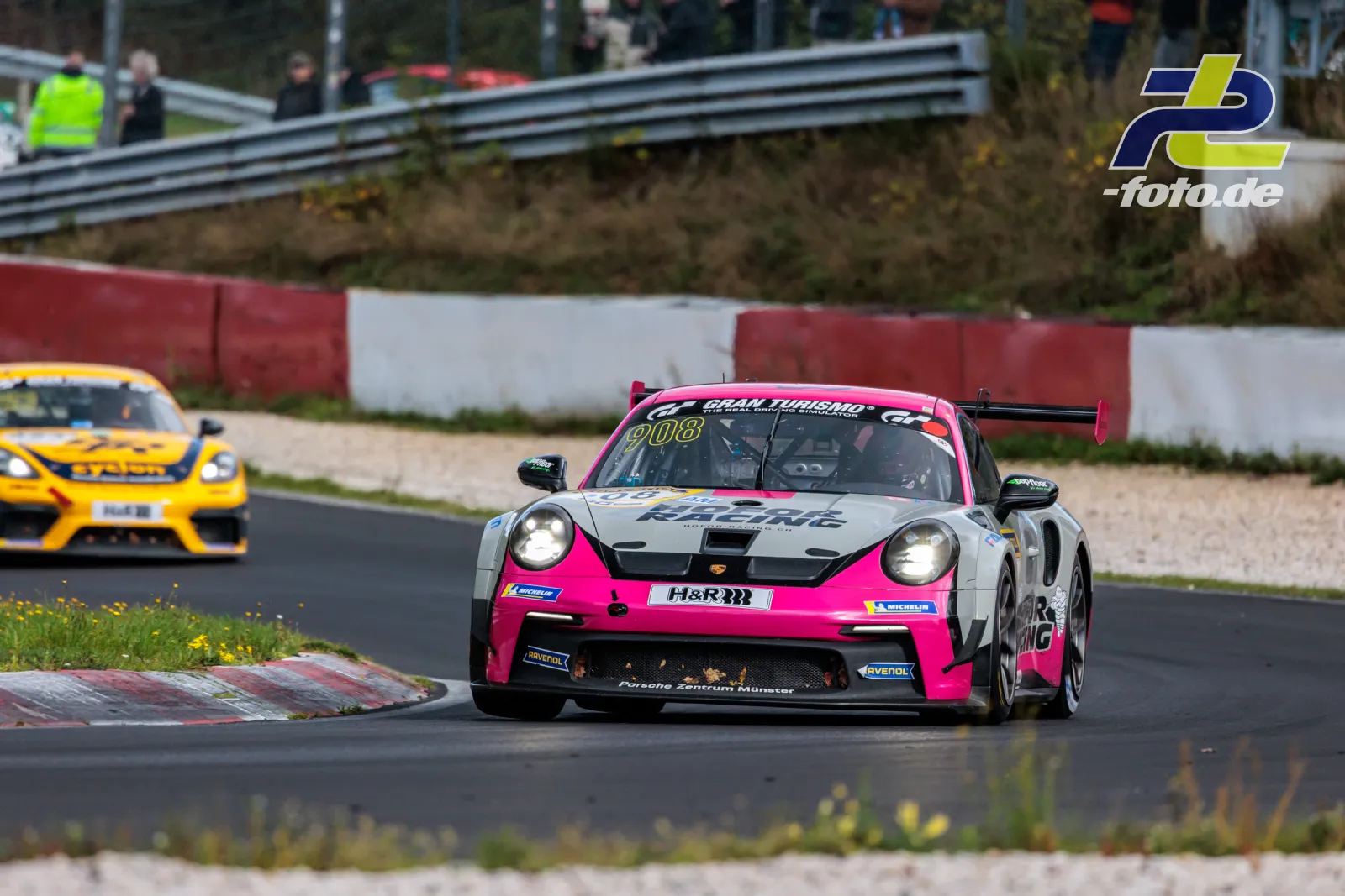 Porsche 911 GT3 Cup #908 in pink-grauer Lackierung auf dem Nürburgring während des NLS-Rennens, gefolgt von einem gelben Fahrzeug.