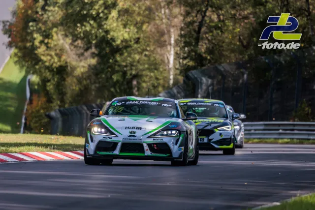 Toyota Supra GT4 in weiß-grüner Lackierung führt ein Feld im Rennen auf dem Nürburgring an, gefolgt von weiteren Fahrzeugen.