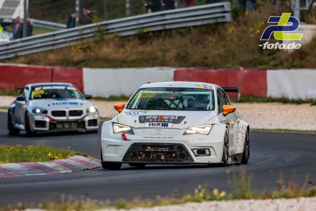 Zwei Rennwagen auf der Nordschleife, vorne ein weißer SEAT mit orangenem Heckflügel, dahinter ein weißer BMW, Randsteinbegrenzung sichtbar.