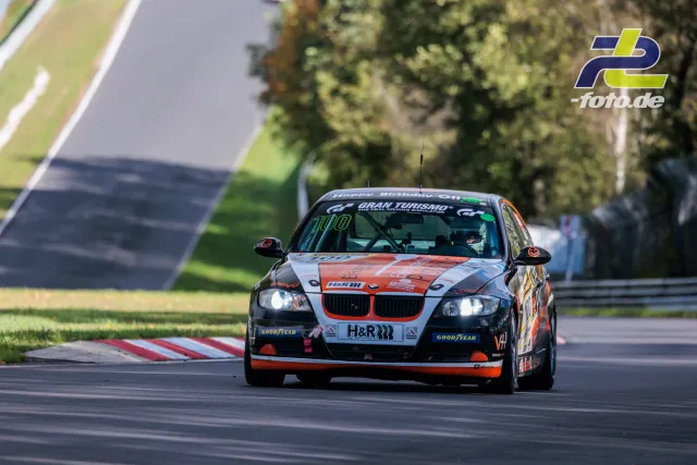 BMW E90 mit silber-oranger Lackierung in einer Kurve auf der Nürburgring-Nordschleife bei Tageslicht mit eingeschalteten Scheinwerfern.