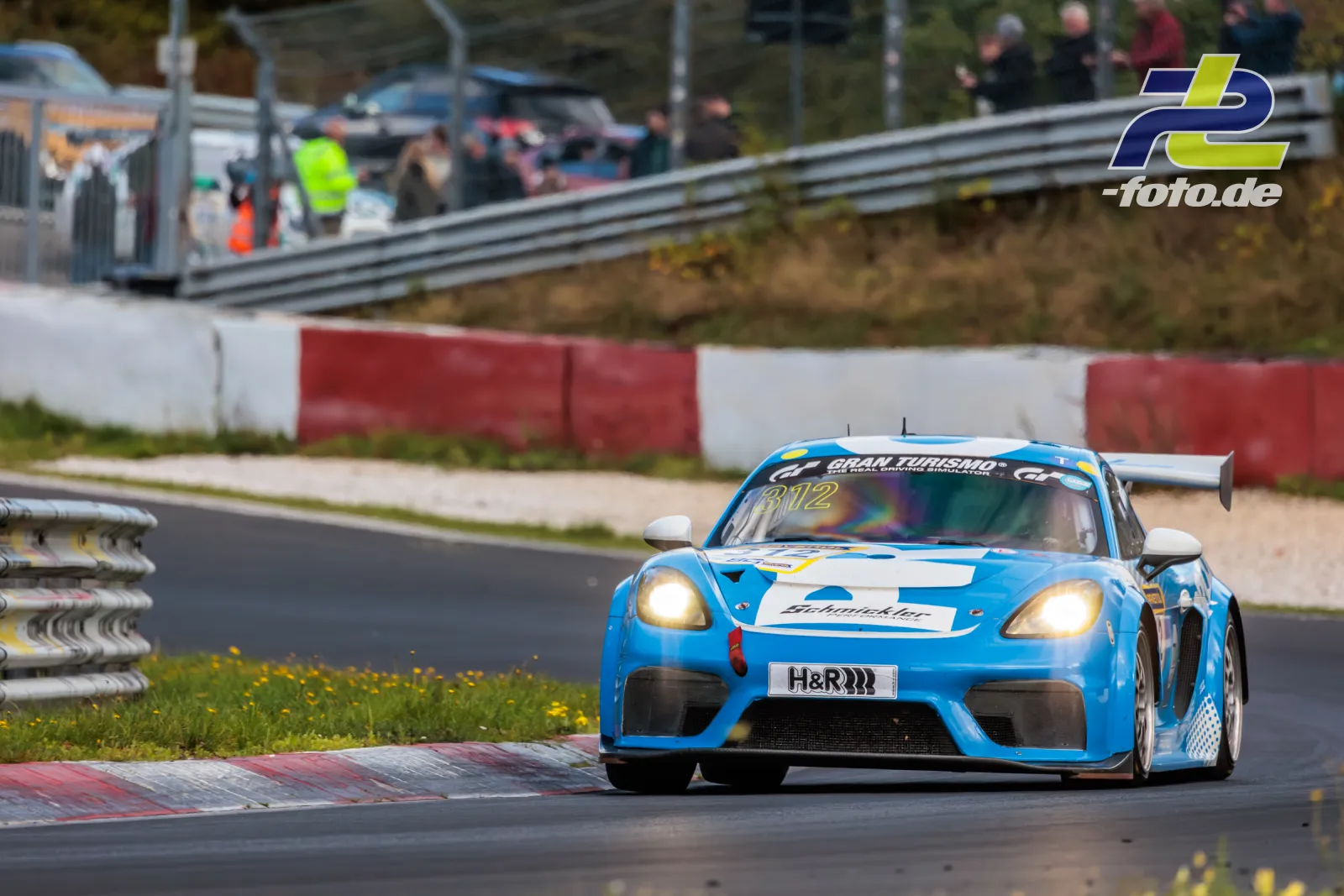 Blau-weißer Porsche 982 Turbo auf der Nordschleife beim NLS-Rennen mit eingeschalteten Scheinwerfern vor Zuschauern.