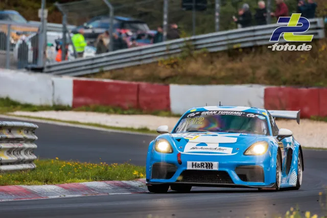 Blauer Porsche 718 Cayman GT4 mit Startnummer 312 in Kurve auf dem Nürburgring, Zuschauer und Leitplanken im Hintergrund