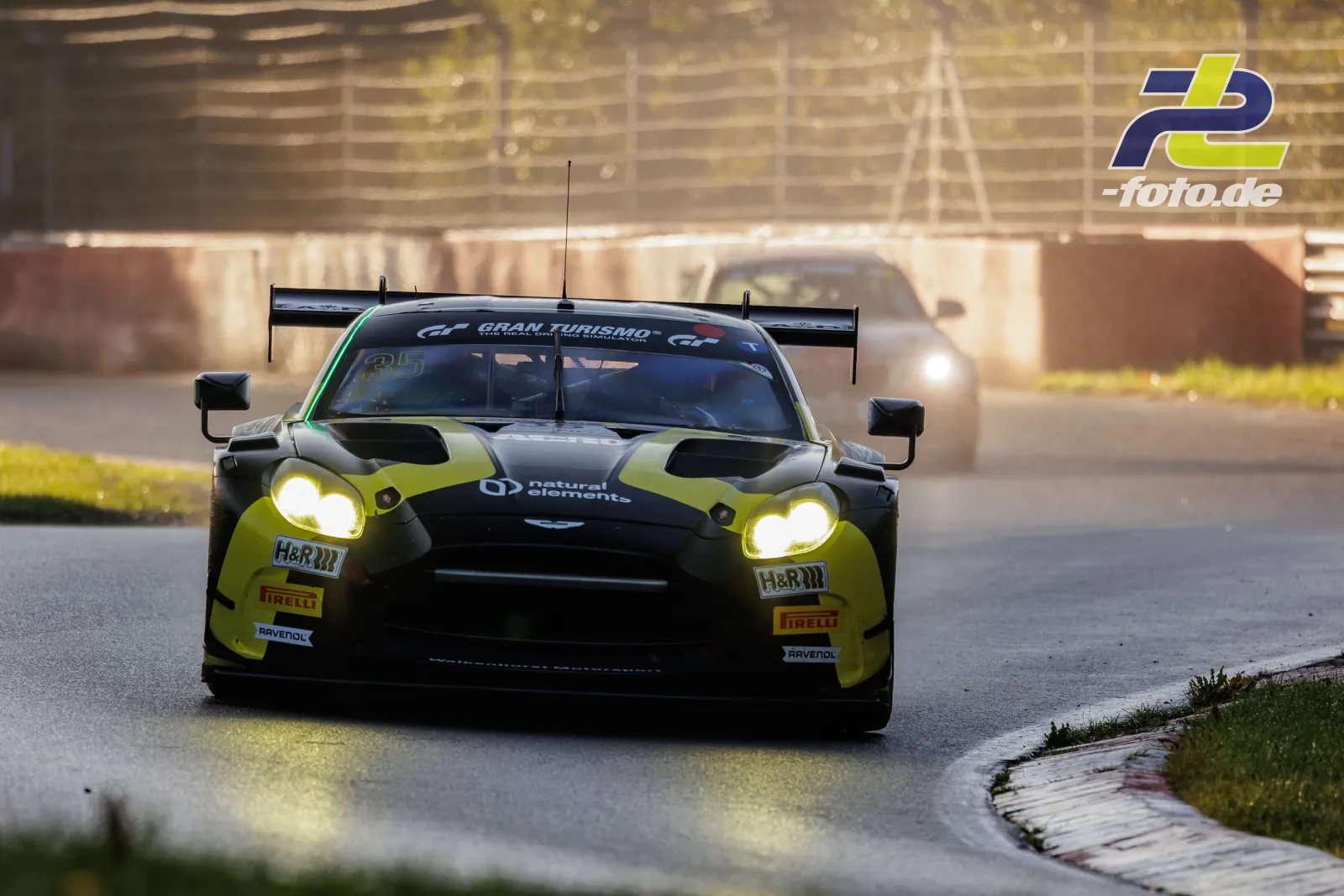 Aston Martin Vantage GT3 EVO in gelb-schwarz im Qualifying der NLS am Nürburgring, Frontansicht in der Kurve mit eingeschalteten Scheinwerfern.