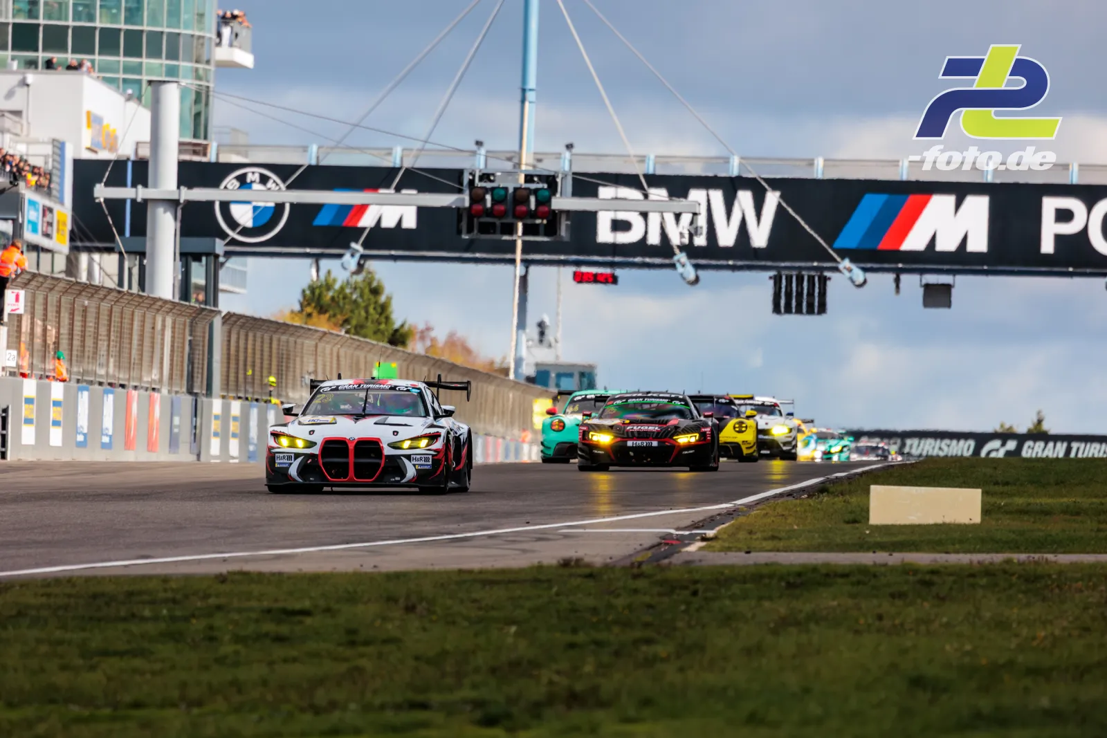GT3-Feld in Boxengasse des Nürburgrings, angeführt von weißem BMW M4 GT3 mit rotem Akzent, Ampel und BMW-Brücke im Hintergrund