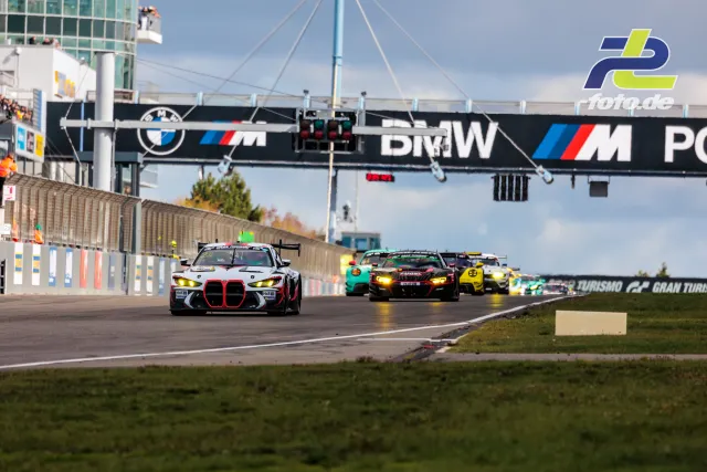 GT3-Feld in Boxengasse des Nürburgrings, angeführt von weißem BMW M4 GT3 mit rotem Akzent, Ampel und BMW-Brücke im Hintergrund
