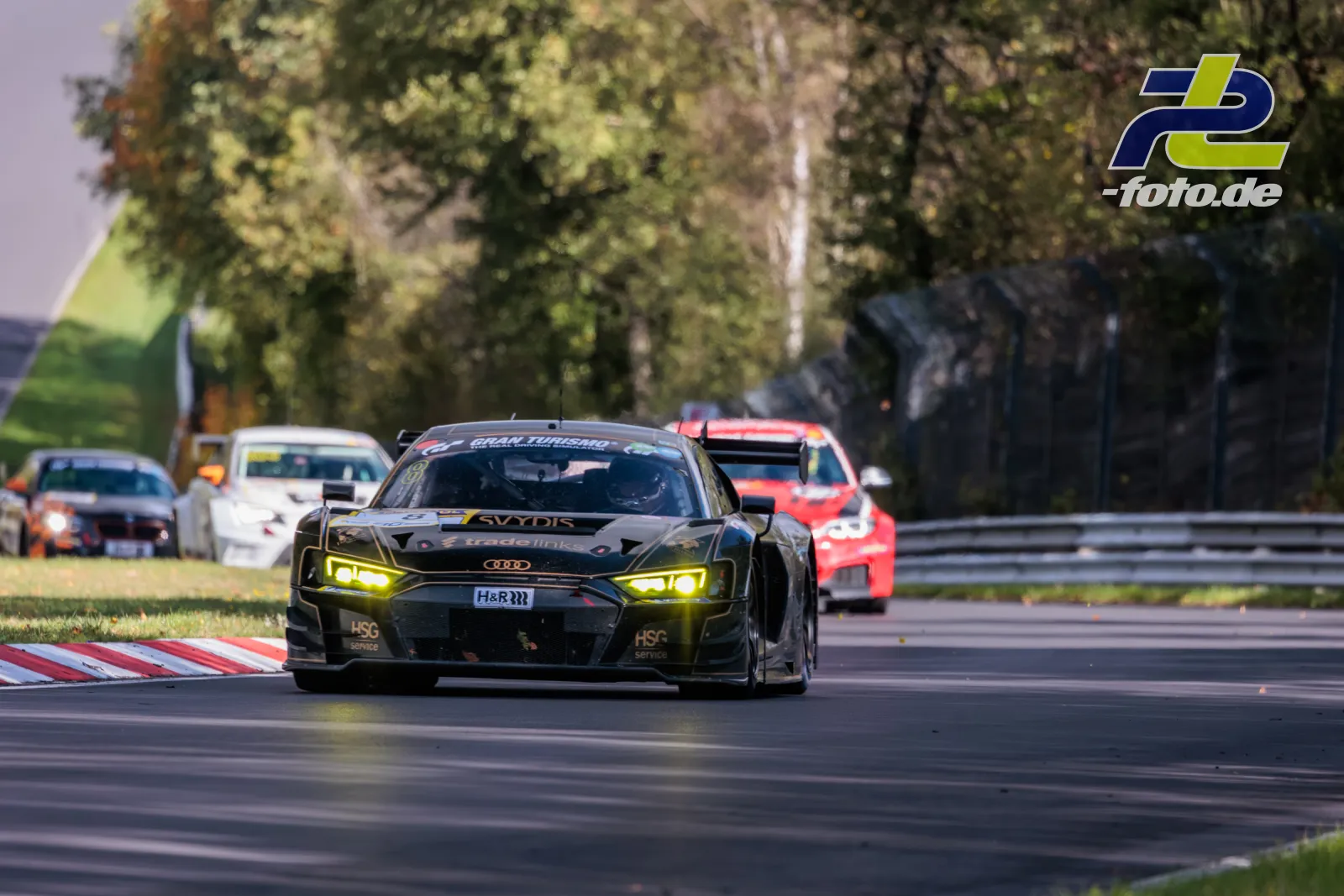 Audi R8 LMS GT3 führt eine Verfolgergruppe auf der Nürburgring-Nordschleife an, gelbe Scheinwerfer leuchten im Sonnenlicht.