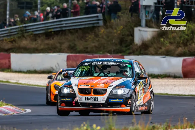 BMW 3er mit Startnummer 100 in orange-blauer Lackierung auf der Nordschleife, gefolgt von zweitem Fahrzeug, Zuschauer im Hintergrund.