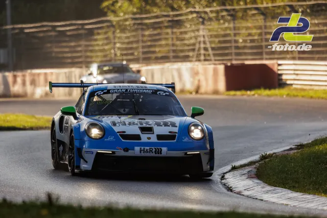 Porsche 911 GT3 R Nr. 904 in blau-weißer Lackierung mit eingeschalteten Scheinwerfern auf nasser Strecke im Gegenlicht, Nürburgring Qualifying.