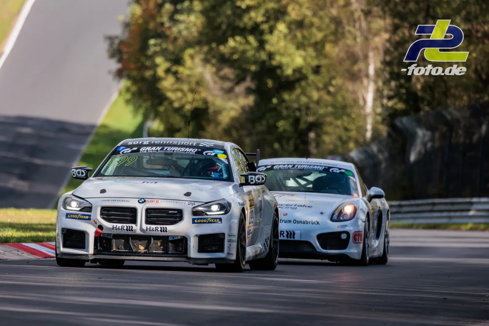 Zwei weiße Rennwagen in Kurve auf dem Nürburgring beim NLS-Rennen, BMW mit Startnummer 99 führend vor Porsche Cayman