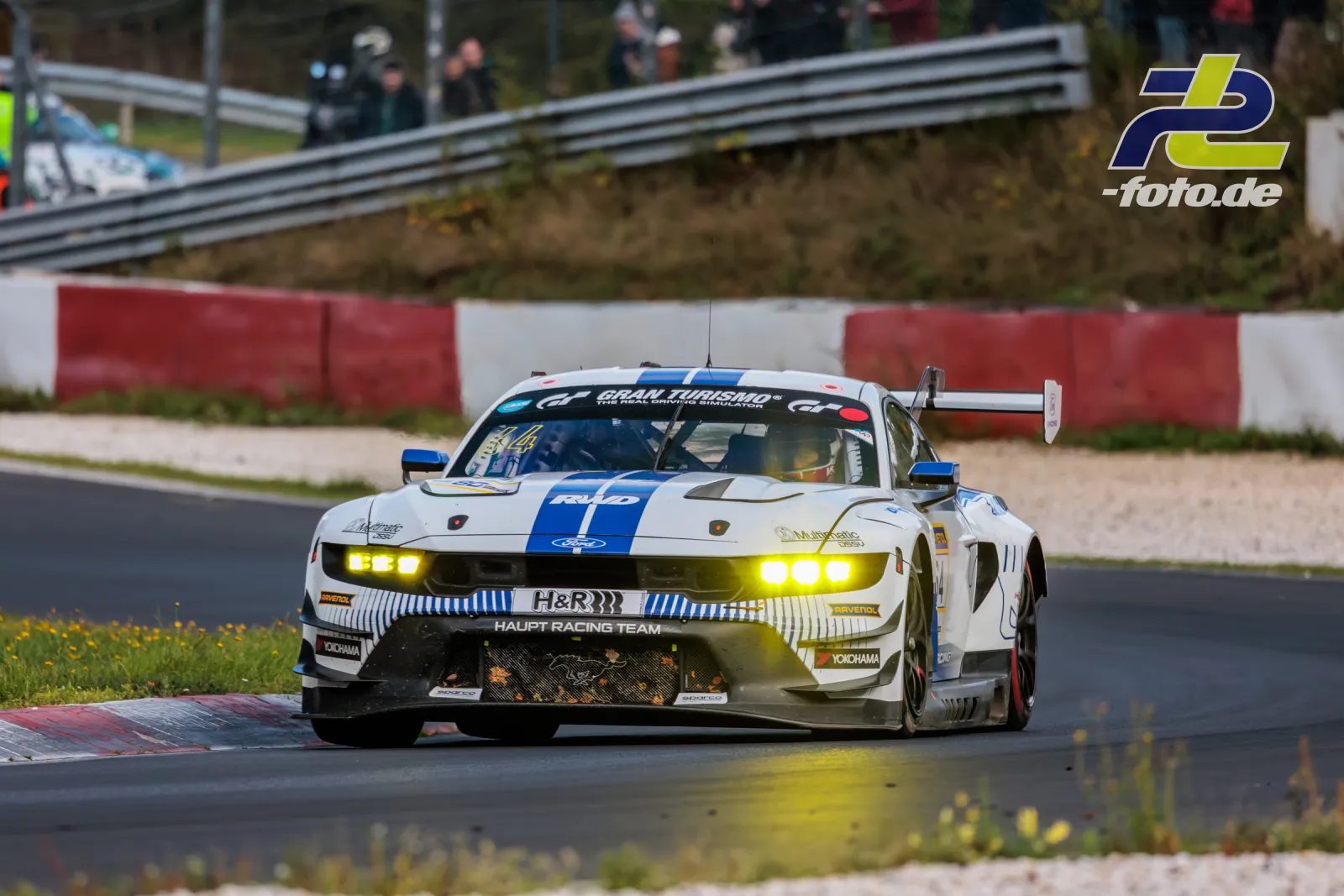 Ford Mustang GT3 des Haupt Racing Team in Kurve am Nürburgring während NLS Lauf 10, Frontansicht mit leuchtenden Scheinwerfern.