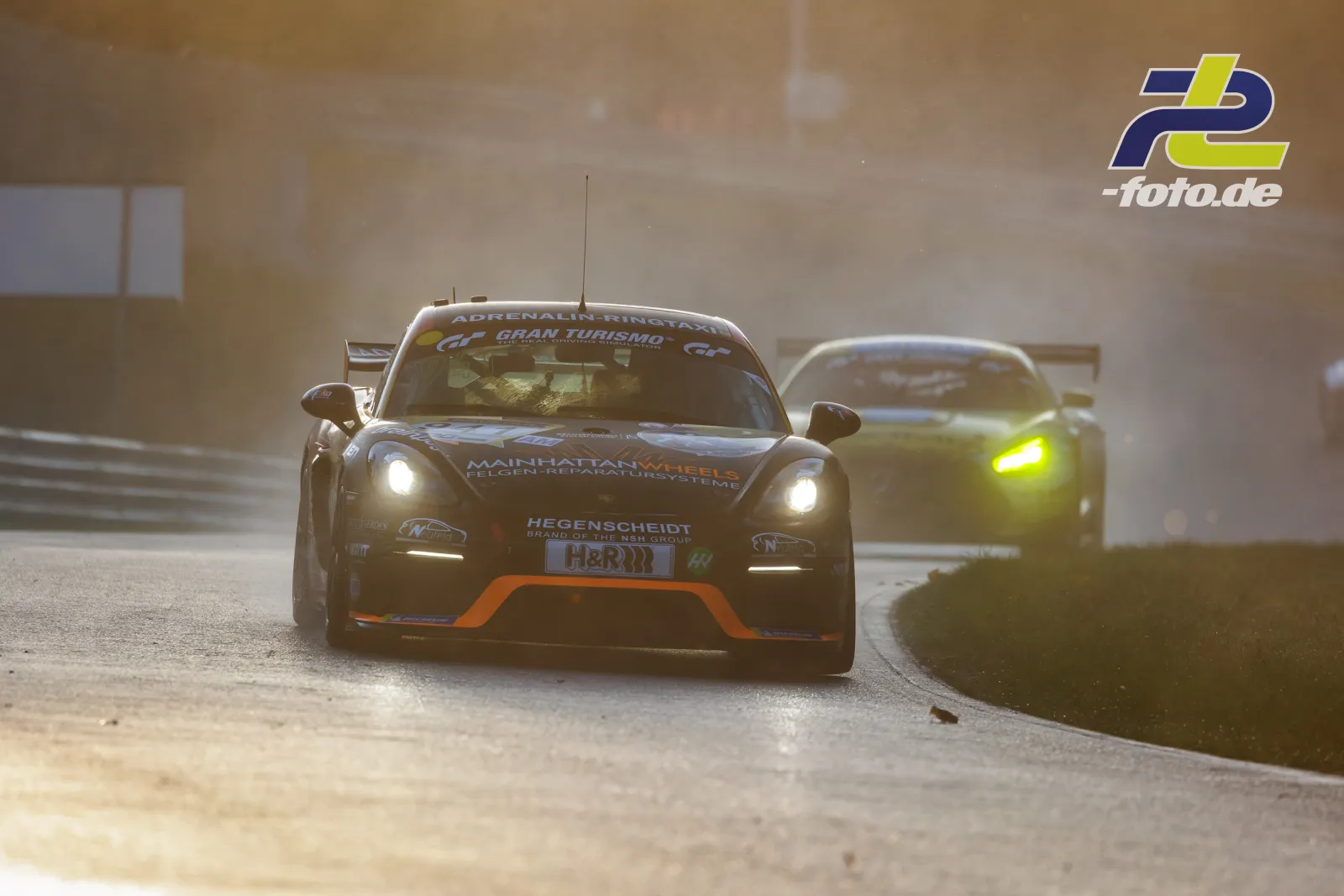 GT-Fahrzeug mit eingeschalteten Scheinwerfern im Gegenlicht auf dem Nürburgring, verfolgt von einem zweiten Sportwagen.