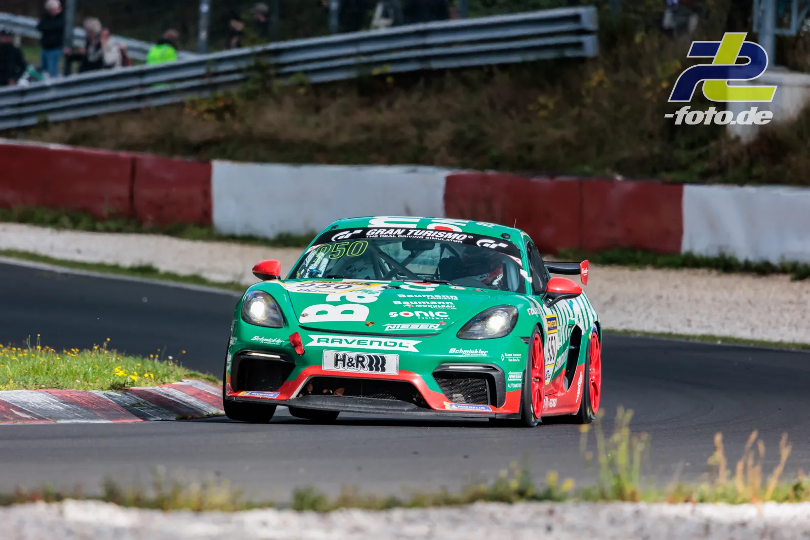 Grün-roter Porsche 718 Cayman GT4 CS #950 in der Kurve beim NLS-Rennen am Nürburgring, fährt über Randstein mit Kiesbett im Hintergrund.