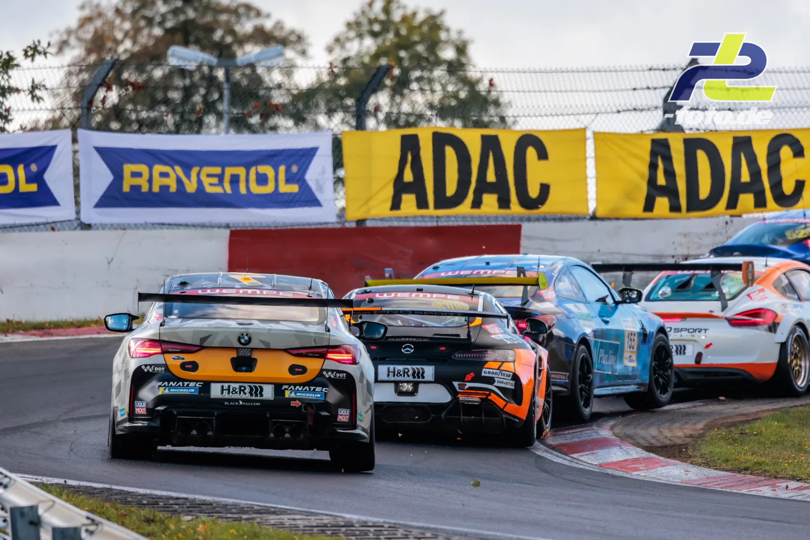 Mehrere GT-Rennwagen fahren durch eine Kurve auf der Nordschleife beim NLS-Rennen, ADAC- und Ravenol-Werbebanden im Hintergrund.