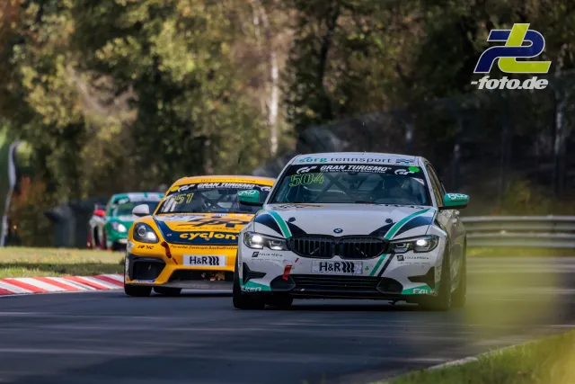 Drei Rennfahrzeuge im Zweikampf auf der Nordschleife, angeführt von einem weißen BMW mit grünen Akzenten vor einem gelben Porsche.