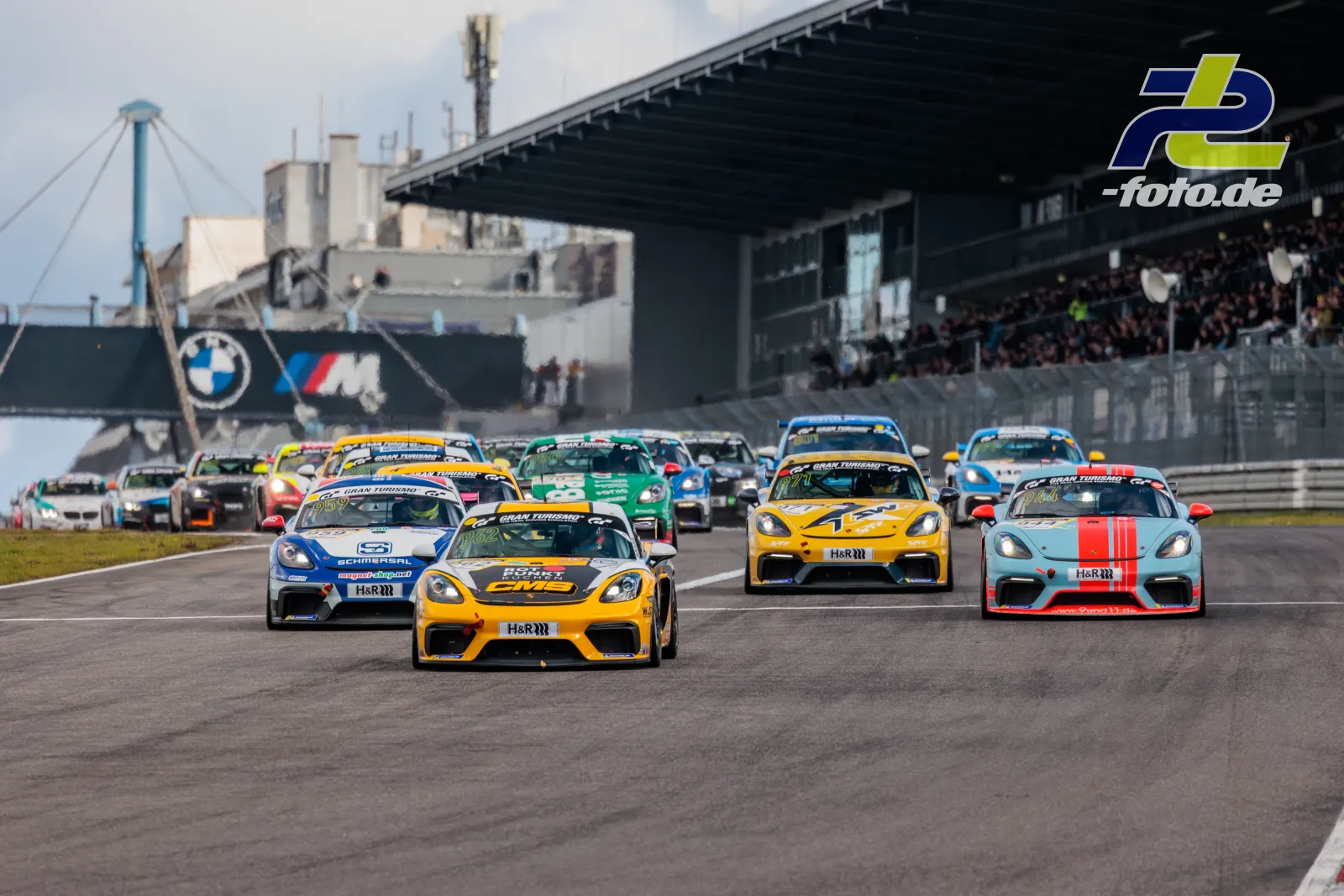 Starterfeld beim NLS-Rennen am Nürburgring mit dicht gepackten Fahrzeugen, Tribüne mit Zuschauern im Hintergrund.