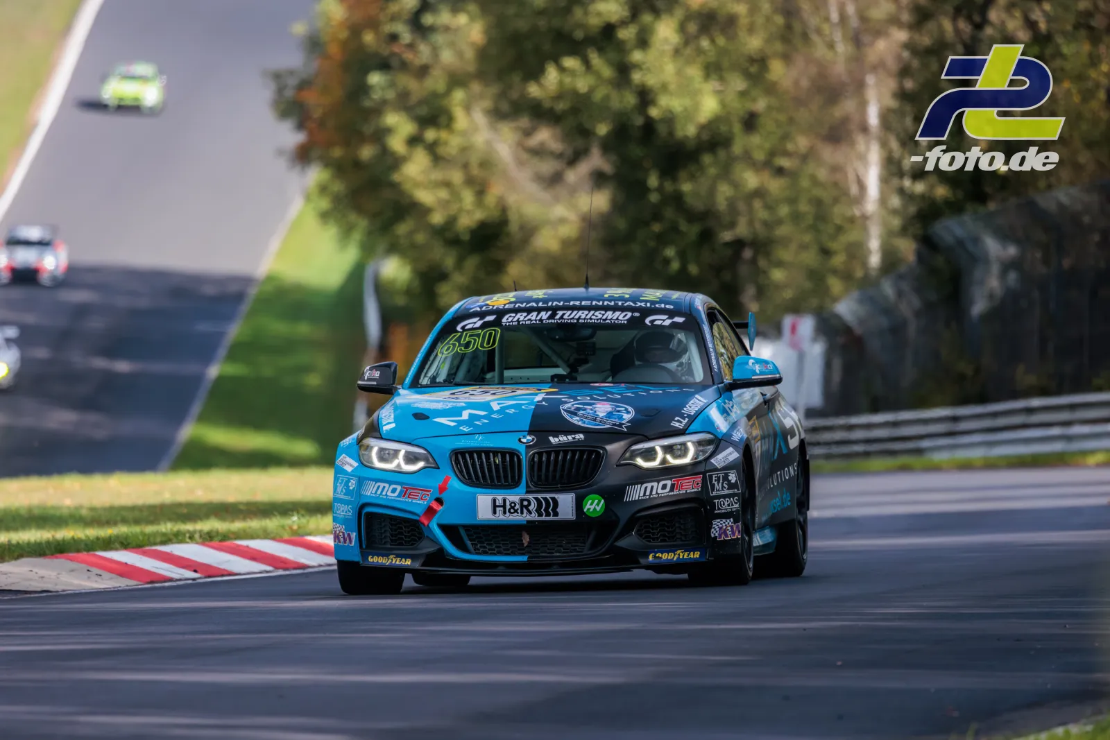 BMW M240i Racing auf der Nürburgring-Nordschleife beim NLS-Rennen, frontal aufgenommen mit Randstein und Grün im Hintergrund.