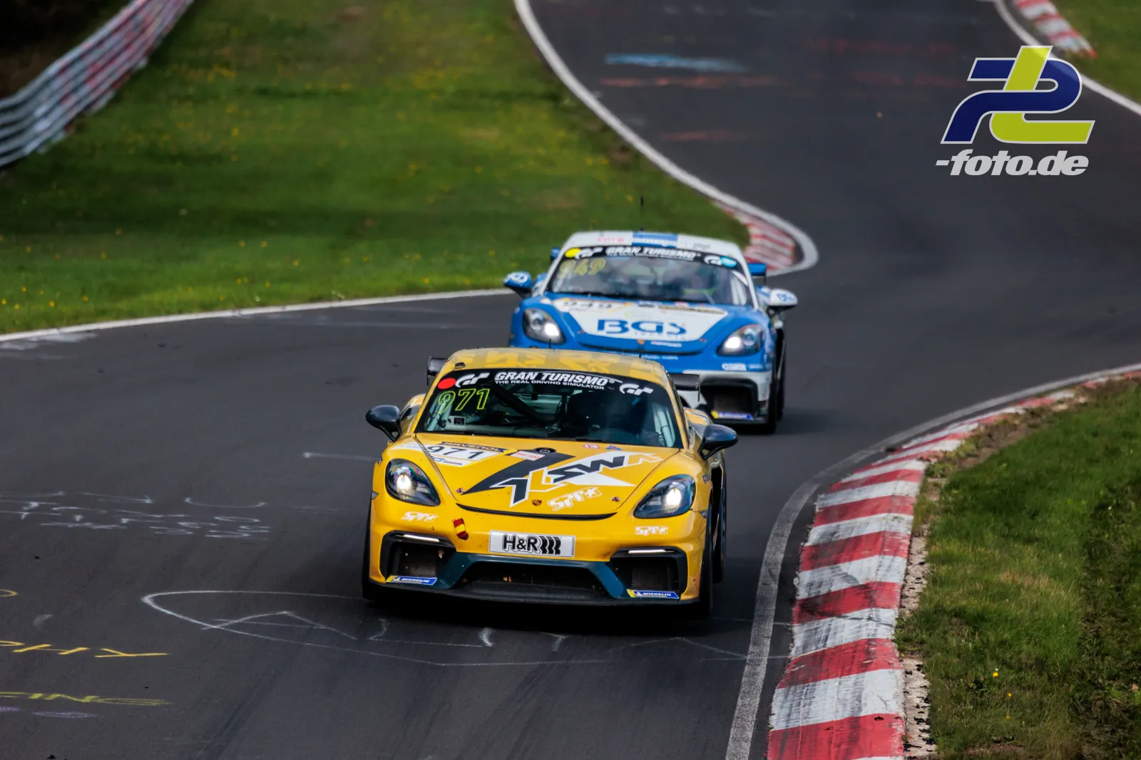 Gelber Porsche 718 Cayman GT4 CS in Kurve auf dem Nürburgring, gefolgt von blauem Fahrzeug während NLS Rennen