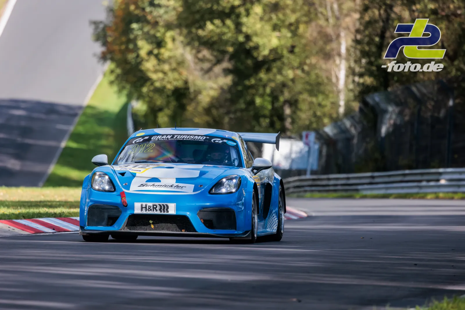 Porsche 982 Turbo in Blau-Weiß auf der Nordschleife beim NLS-Rennen, Frontalansicht in Kurve mit Randsteinen und Bäumen im Hintergrund
