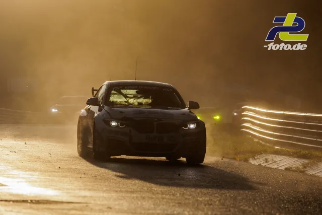 Rennfahrzeug im Gegenlicht auf der Nordschleife bei staubiger Atmosphäre, weitere Fahrzeuge im Hintergrund sichtbar.