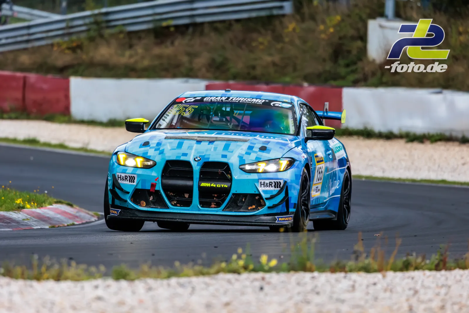 BMW M4 GT4 mit hellblauer Tarnlackierung in Kurve auf dem Nürburgring, Scheinwerfer eingeschaltet, Kiesbett im Vordergrund.
